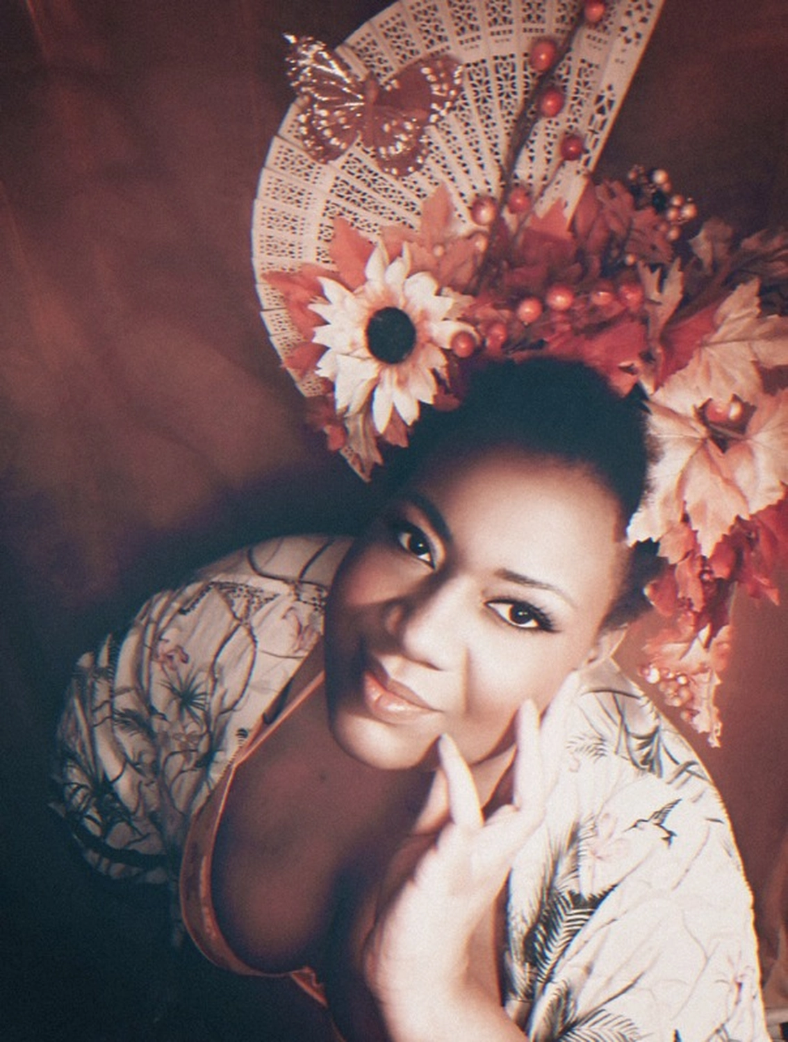 Dela-Noéline Gomado - Zweisprachige Open-Stage-Comedy-Veranstaltung - Noéline, eine Schwarze Frau, trägt einen kunstvollen Kopfschmuck aus herbstlichen Blumen und einen kleinen Holzfächer mit einem Papier-Schmetterling. Sie lächelt schüchtern in die Kamera.